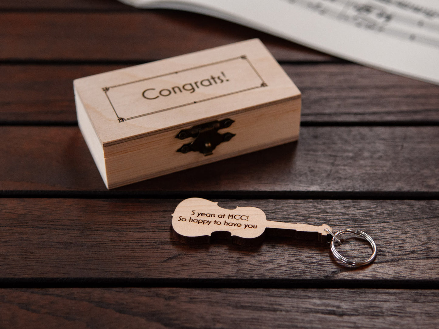 A keychain in the shape of a cello in front of a small wooden box, with custom engraving.