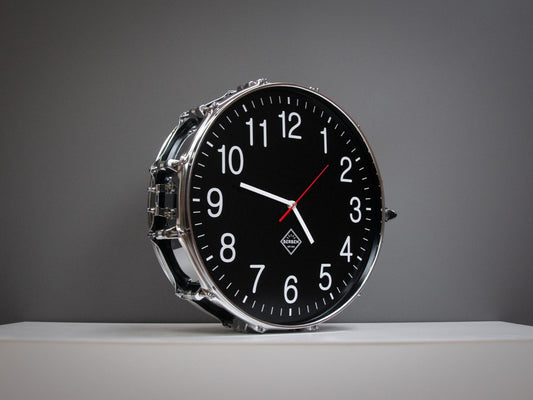 Clock made from a drum with a black clock face, standing on a shelf. The shell and rims have a chrome finish