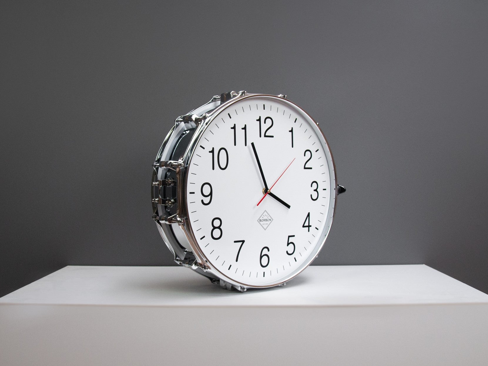 A clock made from a drum with a white clock face, standing on a shelf. The shell and rims have a chrome finish.