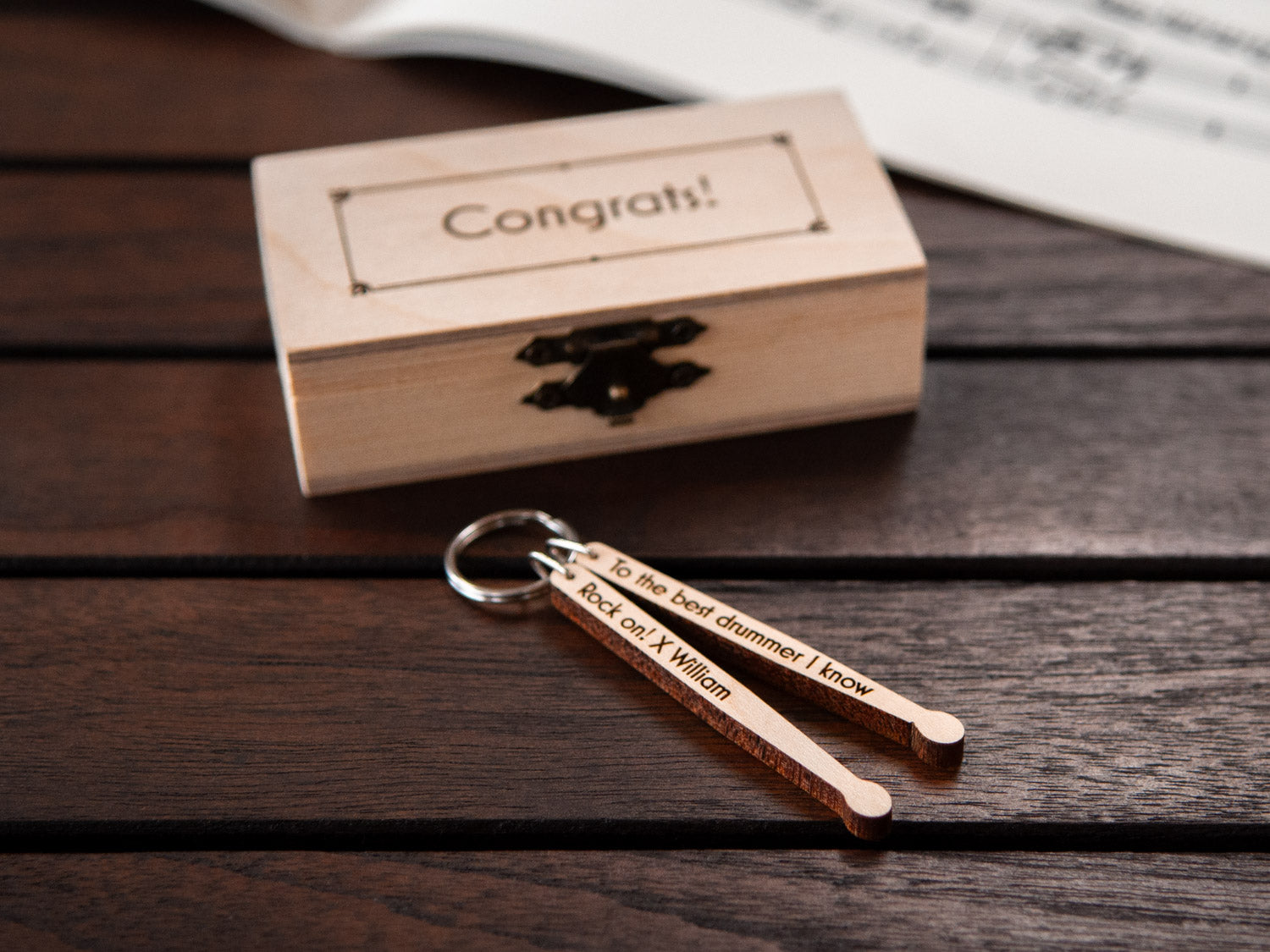 A keychain in the shape of drumsticks in front of a small wooden box, with custom engraving.
