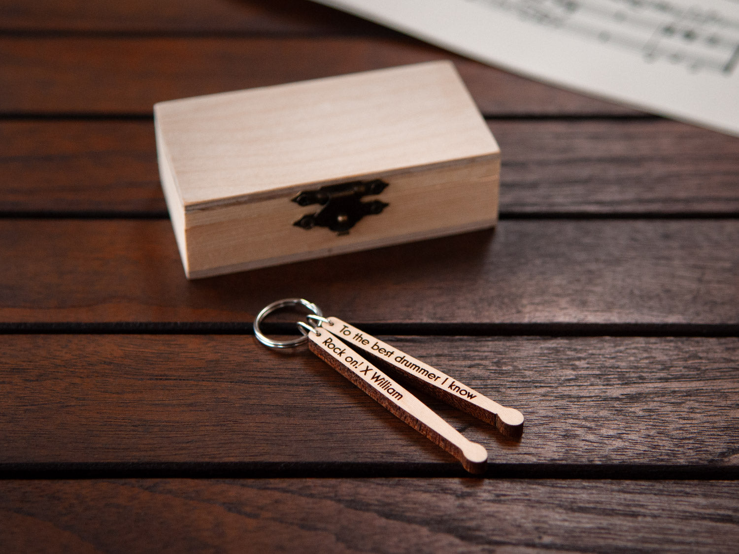 An engraved keychain in the shape drumsticks, in front of a small wooden box