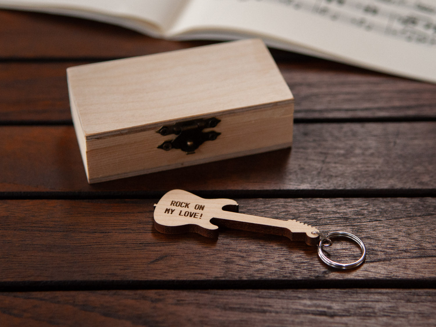 A guitar keychain with personalized engraving positioned in front of a small wooden box.
