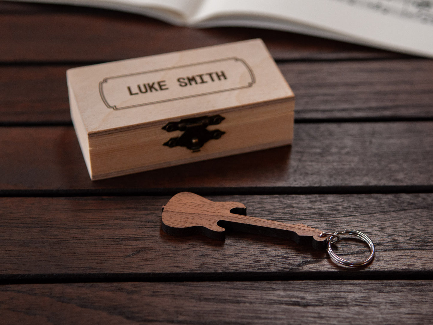 A guitar keychain positioned in front of a small wooden box with engraving on the lid