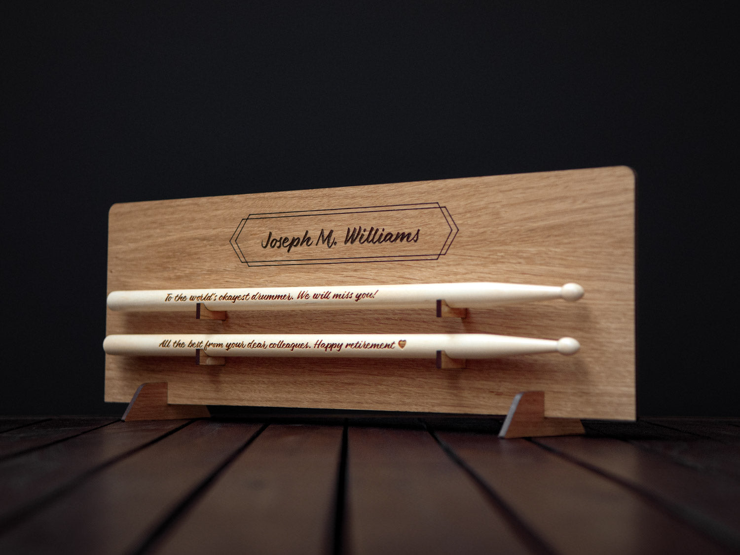 side view of wooden drumsticks with an engraved message on them, placed on a light wooden display with an engraved name on it