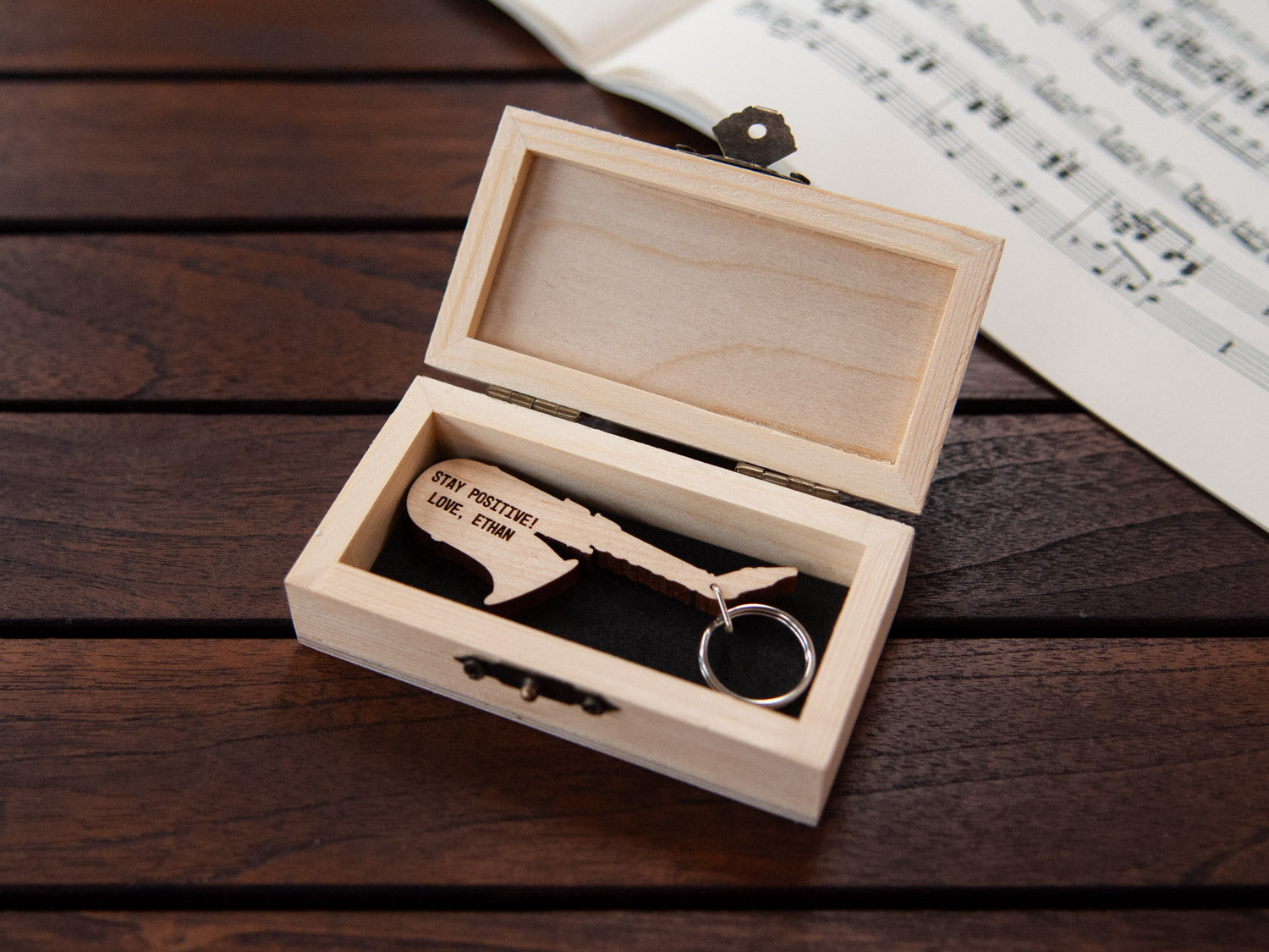 A saxophone shaped keychain placed inside a compact wooden box, featuring a custom engraving.