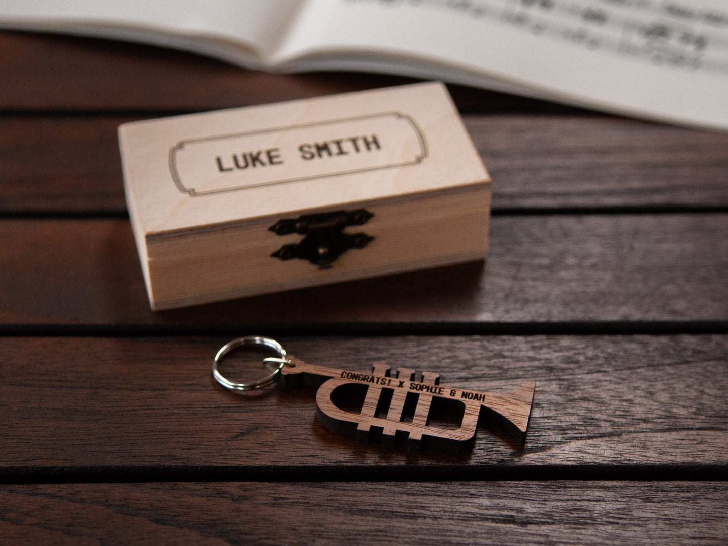 Trumpet keychain displayed in front of a small wooden box, both with a custom engraving.